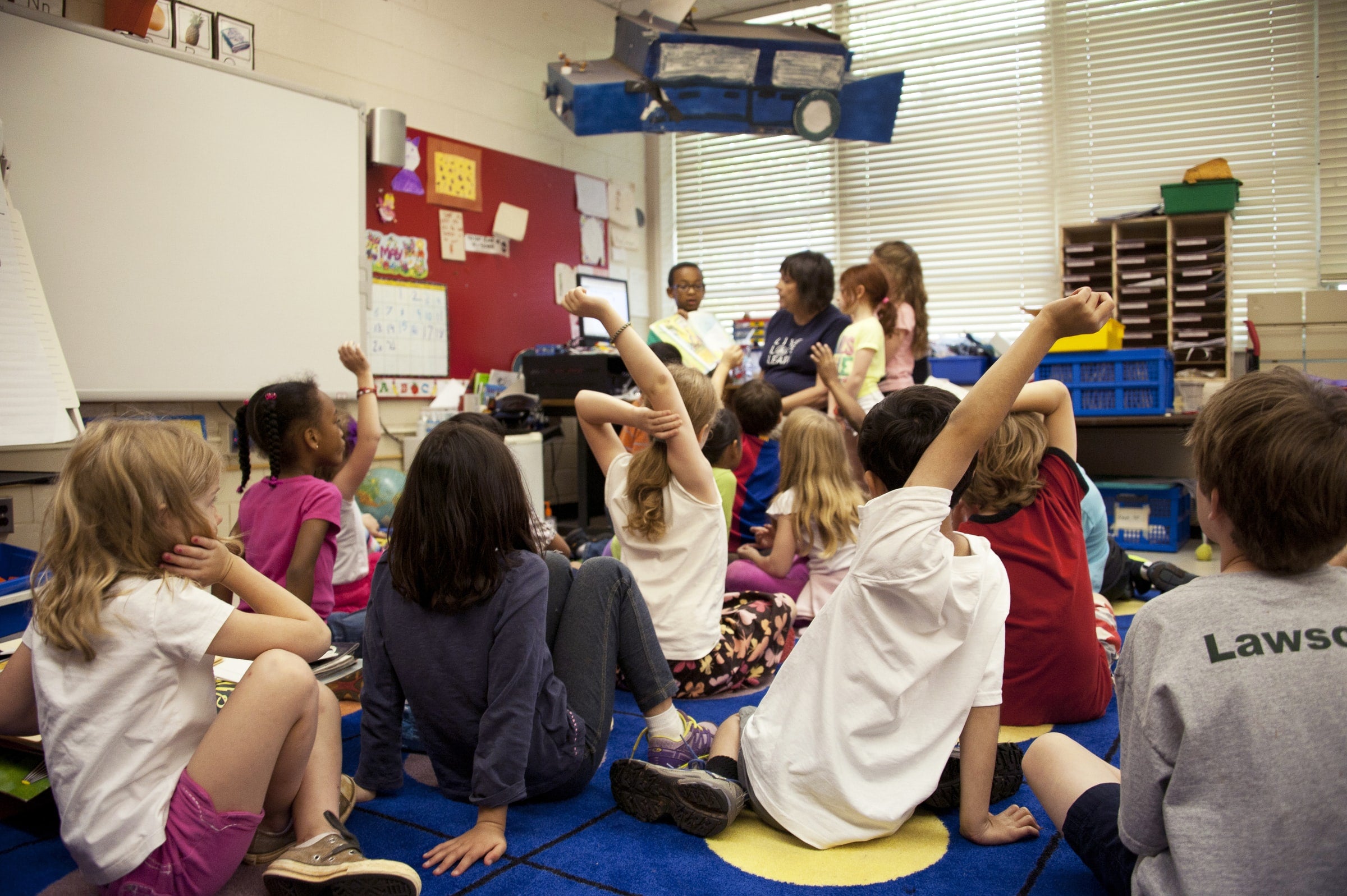 Captured in a metropolitan Atlanta, Georgia primary school, this photograph depicts a typical classroom scene, where an audience of school children were seated on the floor before a teacher at the front of the room, who was reading an illustrated storybook, during one of the scheduled classroom sessions. Assisting the instructor were two female students to her left, and a male student on her right, who was holding up the book, while the seated classmates were raising their hands to answer questions related to the story just read. (Photo/CDC via Unsplash)