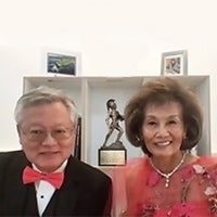 Lunny Ronnie Jung in a white button-down shirt, pink bowtie and black suit jacket and Dianne Kwock in a red dress and necklace with a bookshelf in the background