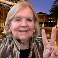 Patsy Dewey in a beige and brown patterned jacket and black shirt holding two fingers up in the USC victory sign