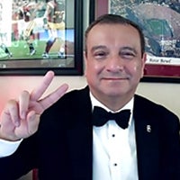 Richard Flores in a white button-down shirt, black bowtie and black suit jacket holding up two fingers in the USC victory sign