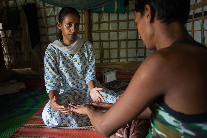 Parveen Parmar in white shirt with a blue-and-yellow pattern conducting a physical exam of a Rohingya man while seated on the floor in a refugee camp building