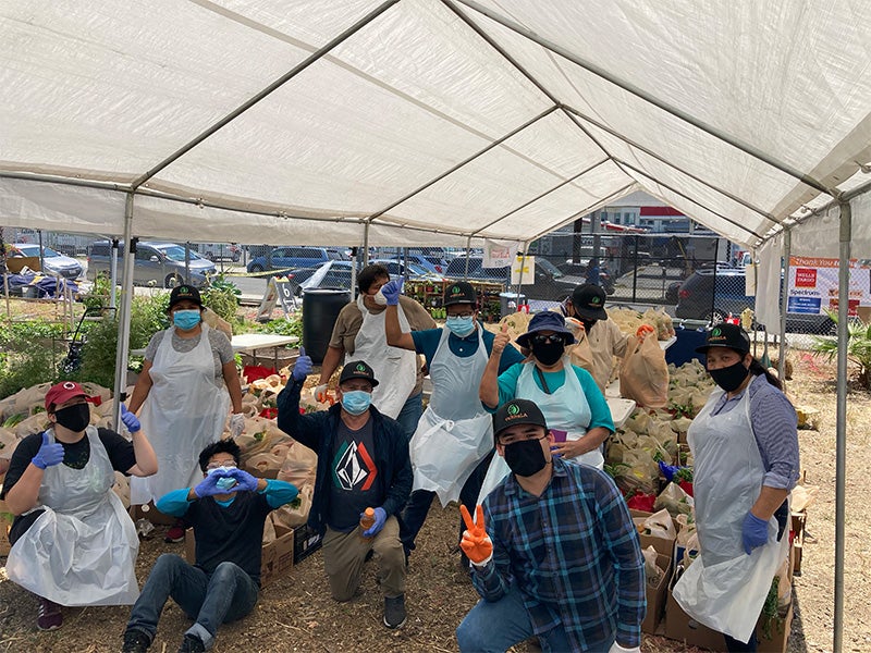 A group of community members in masks, gloves and aprons kneel, sit or stand in front of bags of fresh produce grown in a community garden.