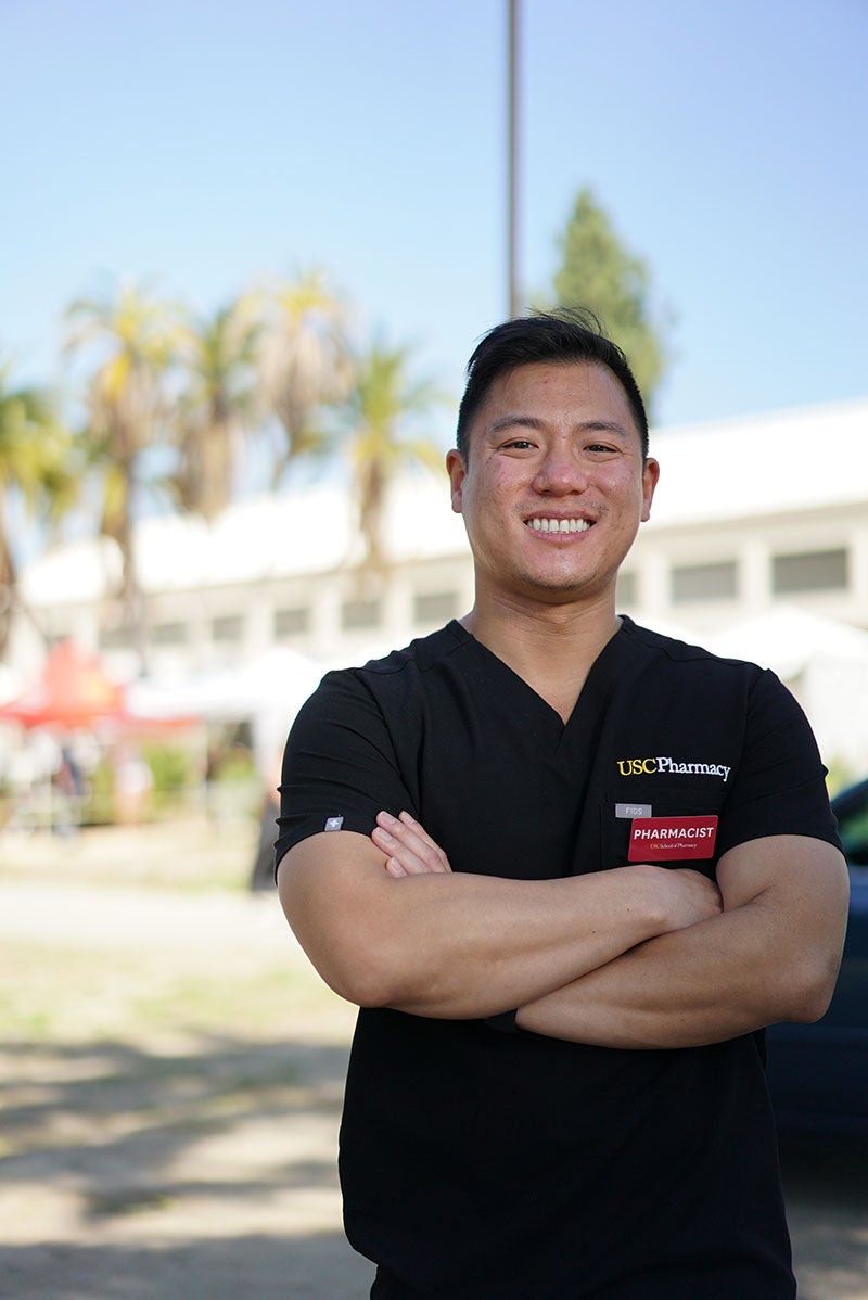 Richard Dang in a black T-Shirt with a USC Pharmacy logo and a red pharmacist badge standing in front of the Lincoln Park vaccination site.