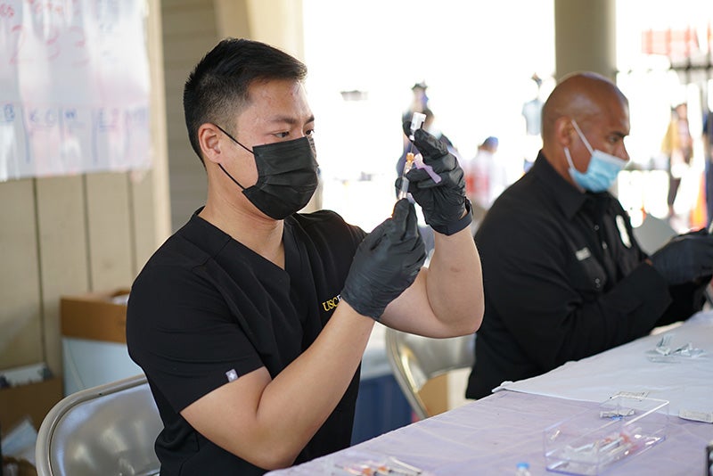 Richard Dang in a black T-shirt, black face mask, and black medical gloves holding a syringe and vial of COVID-19 vaccine.