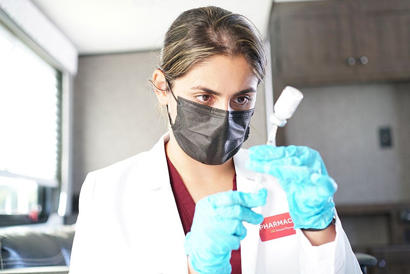 Negin Sazgar in a white lab coat, black face mask and blue medical gloves holds a syringe and vial of COVID-19 vaccine.