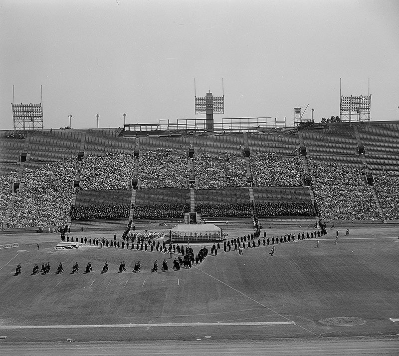 A line of graduates in caps and gowns stretches across the Coliseum field in front of seats filled with onlookers.