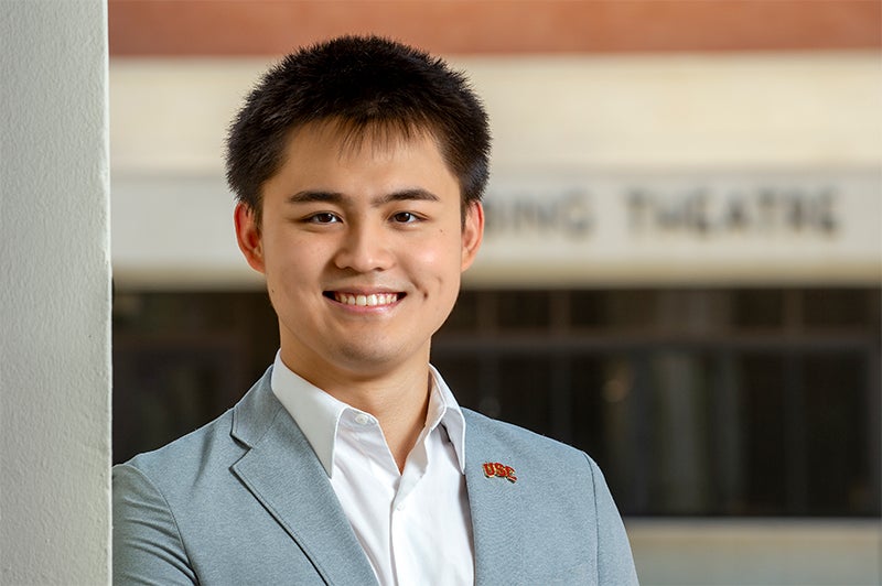 Ziming Jim Liu smiling in a gray suit jacket, white button-down suit and USC lapel pin with Bing Theatre in the background