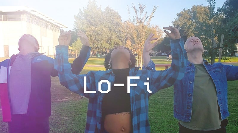 A still from a video showing three people standing in a grassy area with their heads titled back and their arms stretched out to the sides with their hands raised