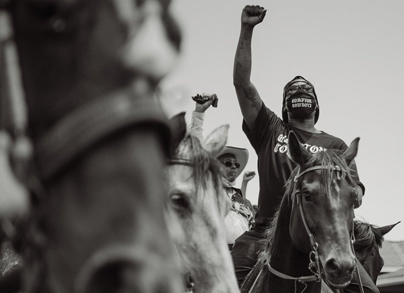 A Black man raising his fist in the air on horseback with a black facemask with the words Compton Cowboys in white type