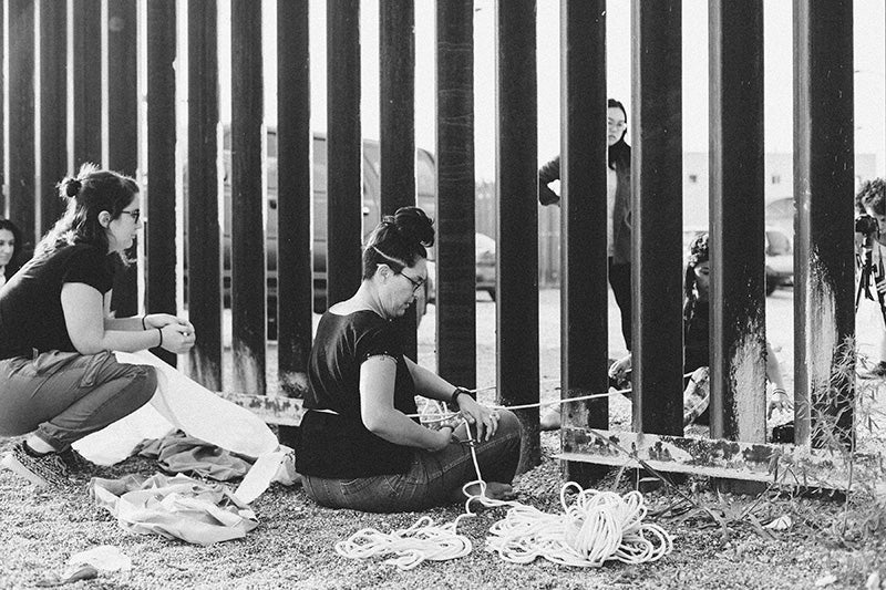 Artist Tanya Aguiñiga seated in front of the U.S.-Mexico border fence with string and other material on the ground around her