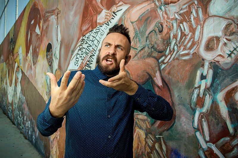 David A. Romero in a blue shirt with white dots gestures with upraised palms while standing against a wall painted with a mural