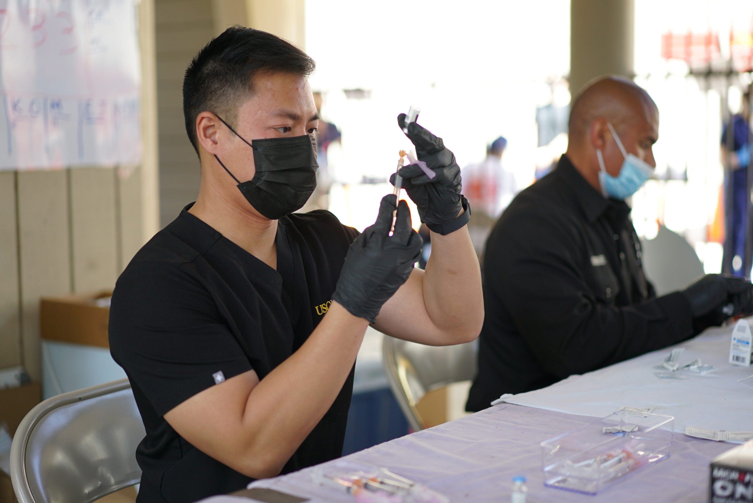 Richard Dang seated at a table in black medical scrubs, gloves and a mask filling a syringe from a vial of COVID-19 vaccine