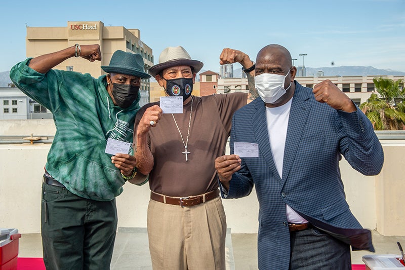 Arsenio Hall, Danny Trejo, and Magic Johnson hold up their COVID-19 vaccine cards and flex their biceps on a rooftop with with downtown Los Angeles in the background