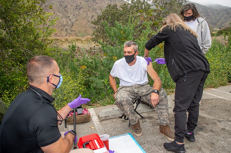 A health worker in black clothing and purple gloves delivering a vaccine shot to the arm of a man in a white T-shirt and camouflage pants seated on a parking curb