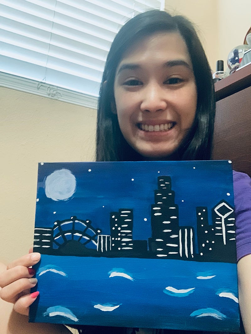 Whitney Tang displaying a blue painting of a skyline at night with water in the foreground