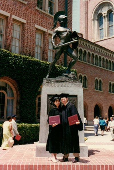 Joyce and Craig Rohan in graduation robes and hats holding USC degree booklets in front of Tommy Trojan.