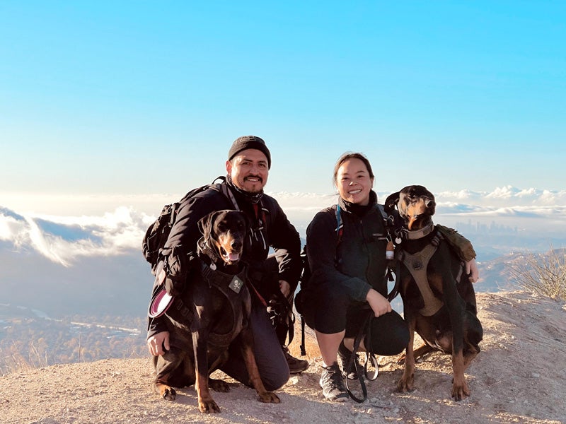 Dahliena and Bryan Chavac on a mountaintop overlooking the Los Angeles skyline with their two Doberman Pinscher dogs.