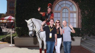 Three people in USC outfits posing with Tommy Trojan and Traveler