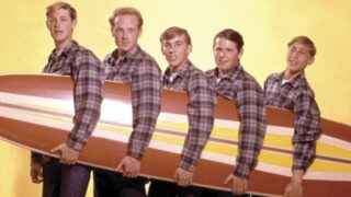 The Beach Boys wearing matching outfits holding a surfboard together
