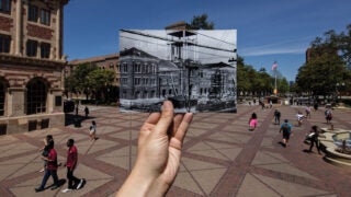 USC Bovard Auditorium in the present with an hand holding a picture up of the same location in the past during construction