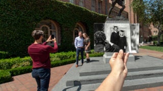 Family posing with son in front of Tommy Trojan as someone holds up an old photo of a family in the past doing the same thing.