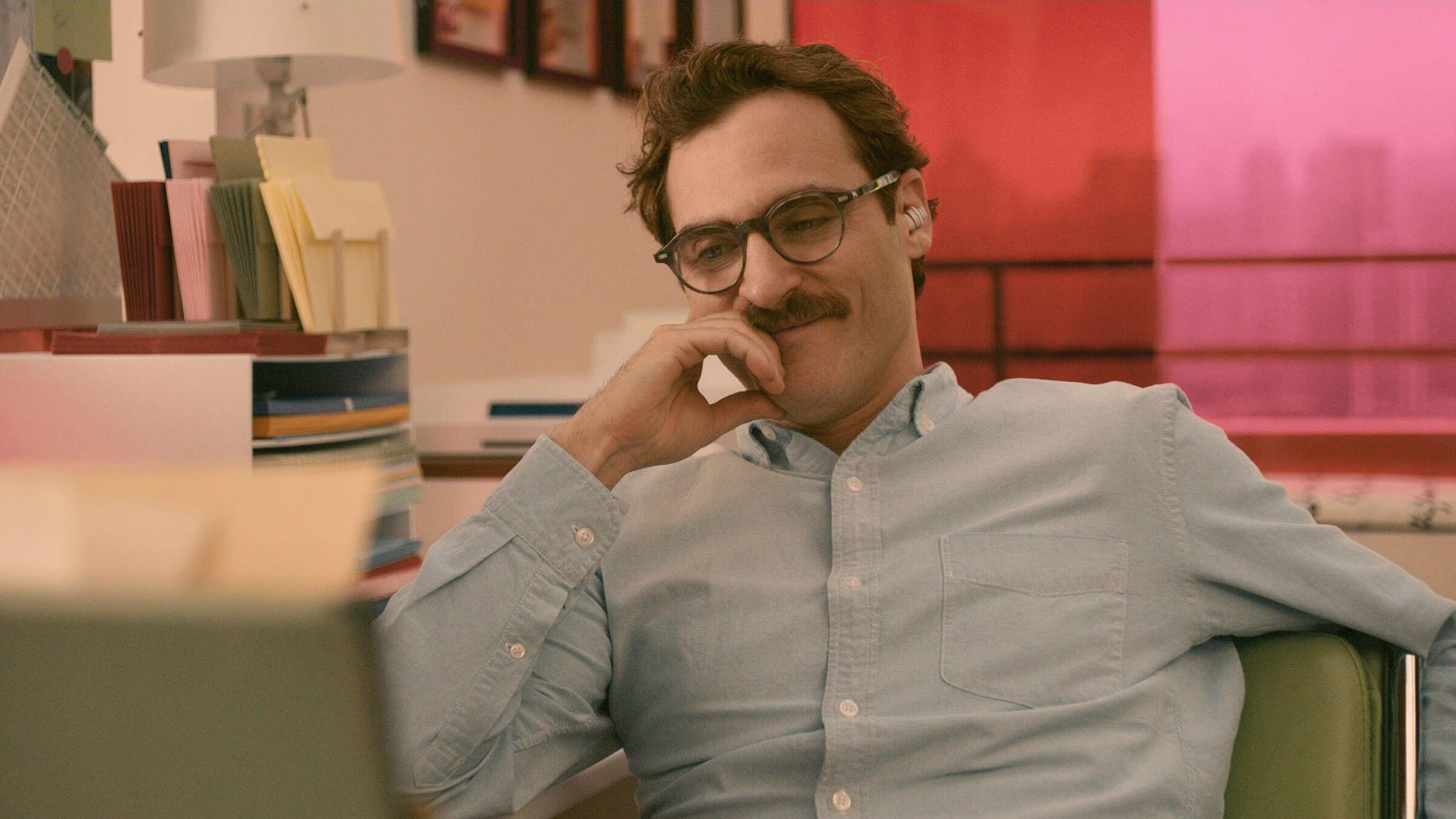 Joaquin Phoenix in a blue shirt in glasses sitting down looking at a monitor.