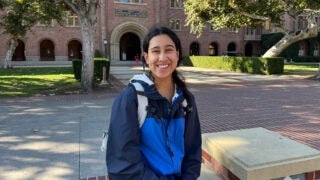 Solana Marquez, a transfer student, smiling in a blue jacket at USC with a brick building and trees in the background.