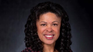 Stayce Harris, smiling in a red and black top against a dark background.