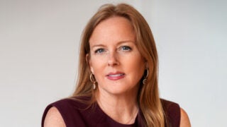 Reon Roski, wearing a burgundy top and gold earrings, against a plain background.