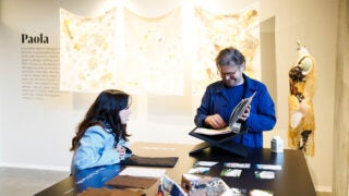 Paola Espinosa and Brian O’Connell at a table with fabric samples and a book, near a dress form and textile art display.