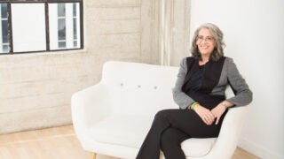 Elyn Saks, wearing glasses and a black suit with a patterned jacket, sitting on a white couch.