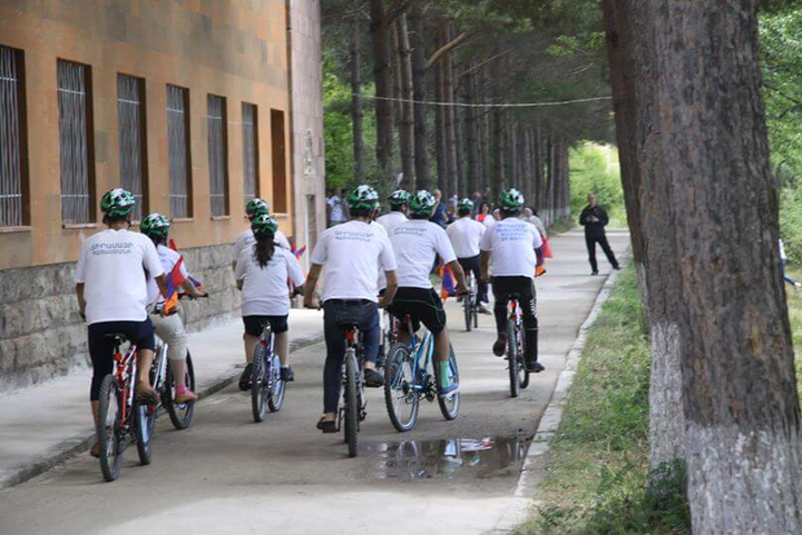 A group of people riding bikes on a path through trees and an orange stone building in Armenia, all in matching green helmets and white t shirts.
