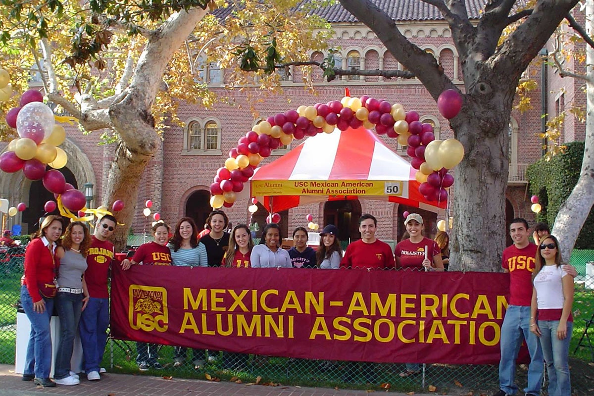 USC alumni celebrating the Mexican American Alumni Association with a sign, standing outside Bovard.