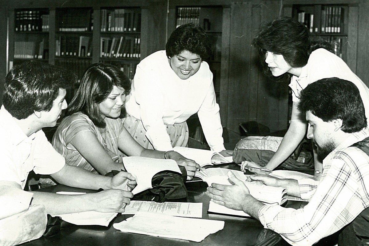 A black and white photo of students around a table looking at books.