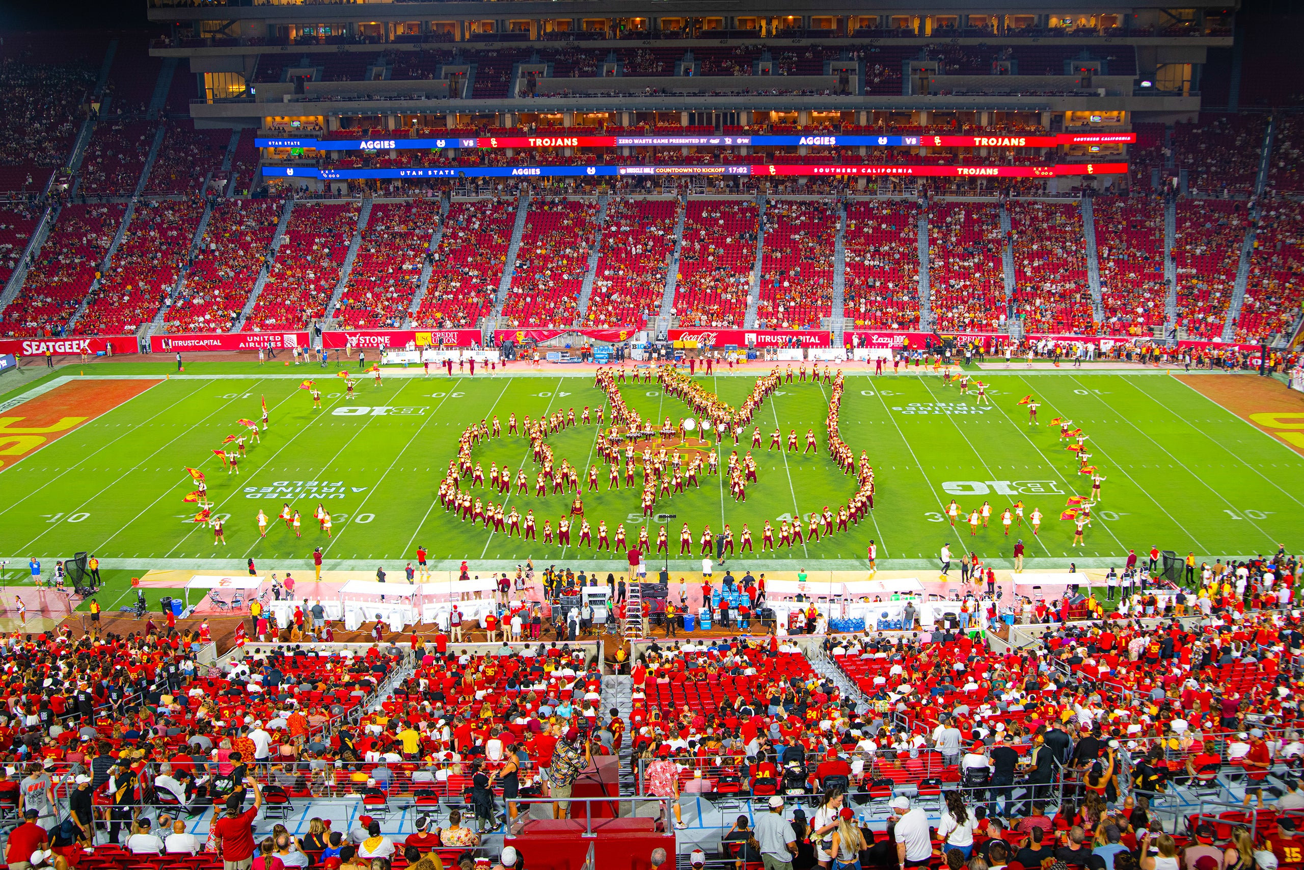 USC marching band forms a large Fight On hand symbol at the USC Coliseum, with cheerleaders and a crowd of spectators in a packed stadium.