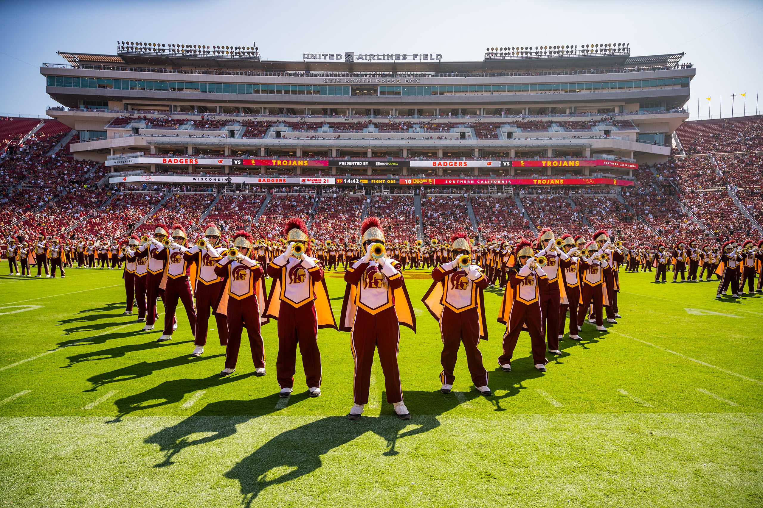 USC marching band in gold and red uniforms performs on the field at Coliseum, with a large stadium filled with spectators in the background.