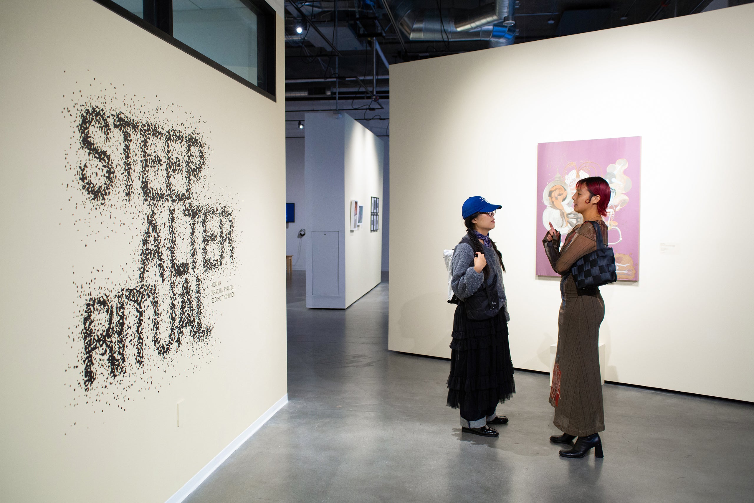 Two people in an art gallery discussing a painting, with 'STEEP ALTER RITUAL' wall text and industrial ceiling.
