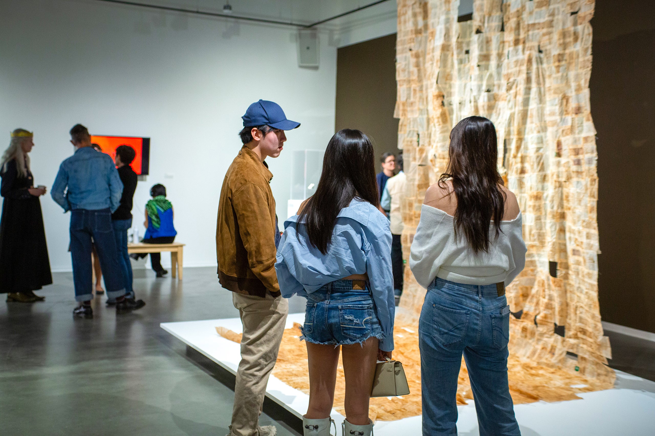 Three youths view an art installation in a modern gallery with paper strip artwork and other visitors in the background.