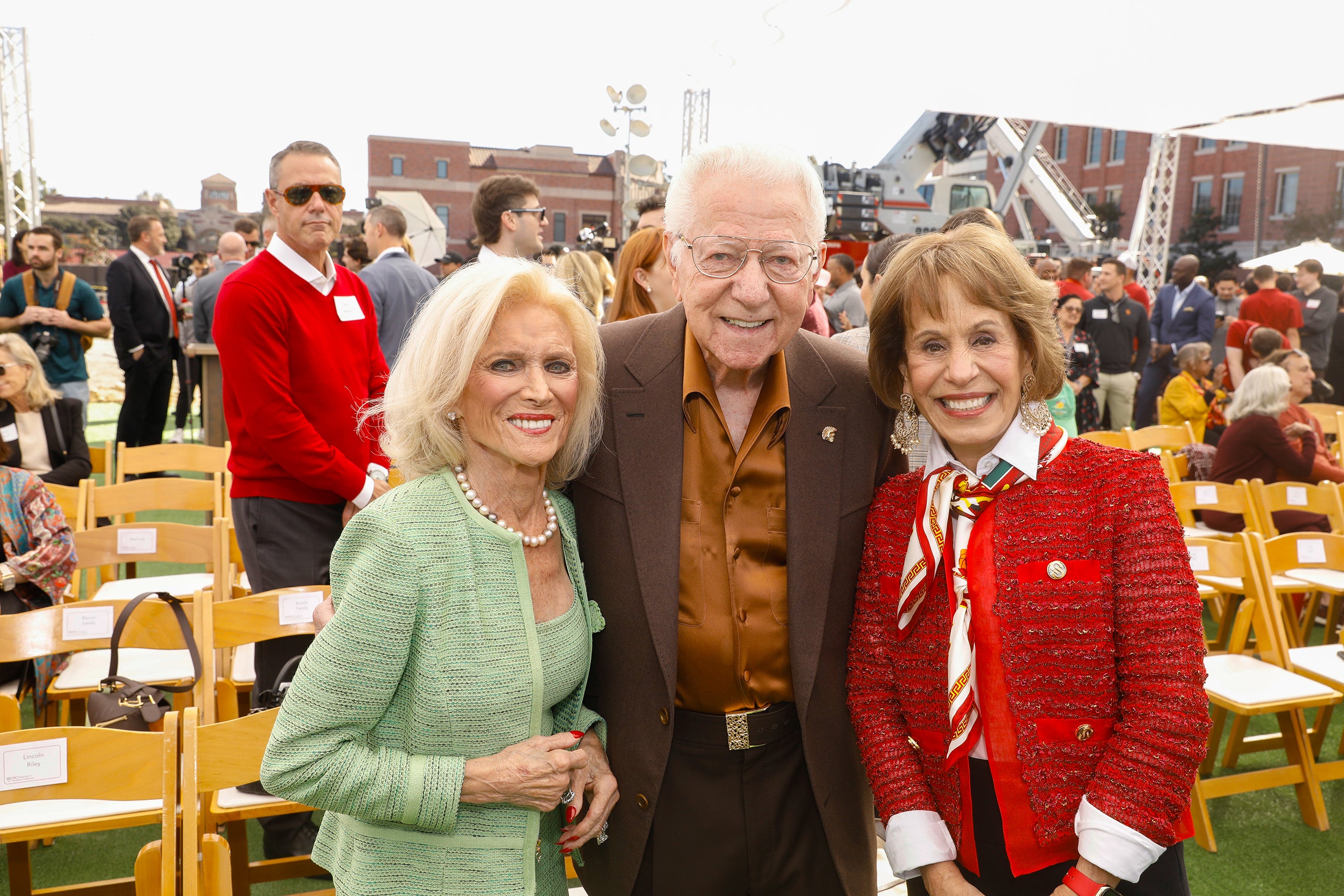 Louis Bloom, Ronald Bloom and President Carol Folt at an outdoor event, wearing colorful attire, with chairs and a crowd behind them.