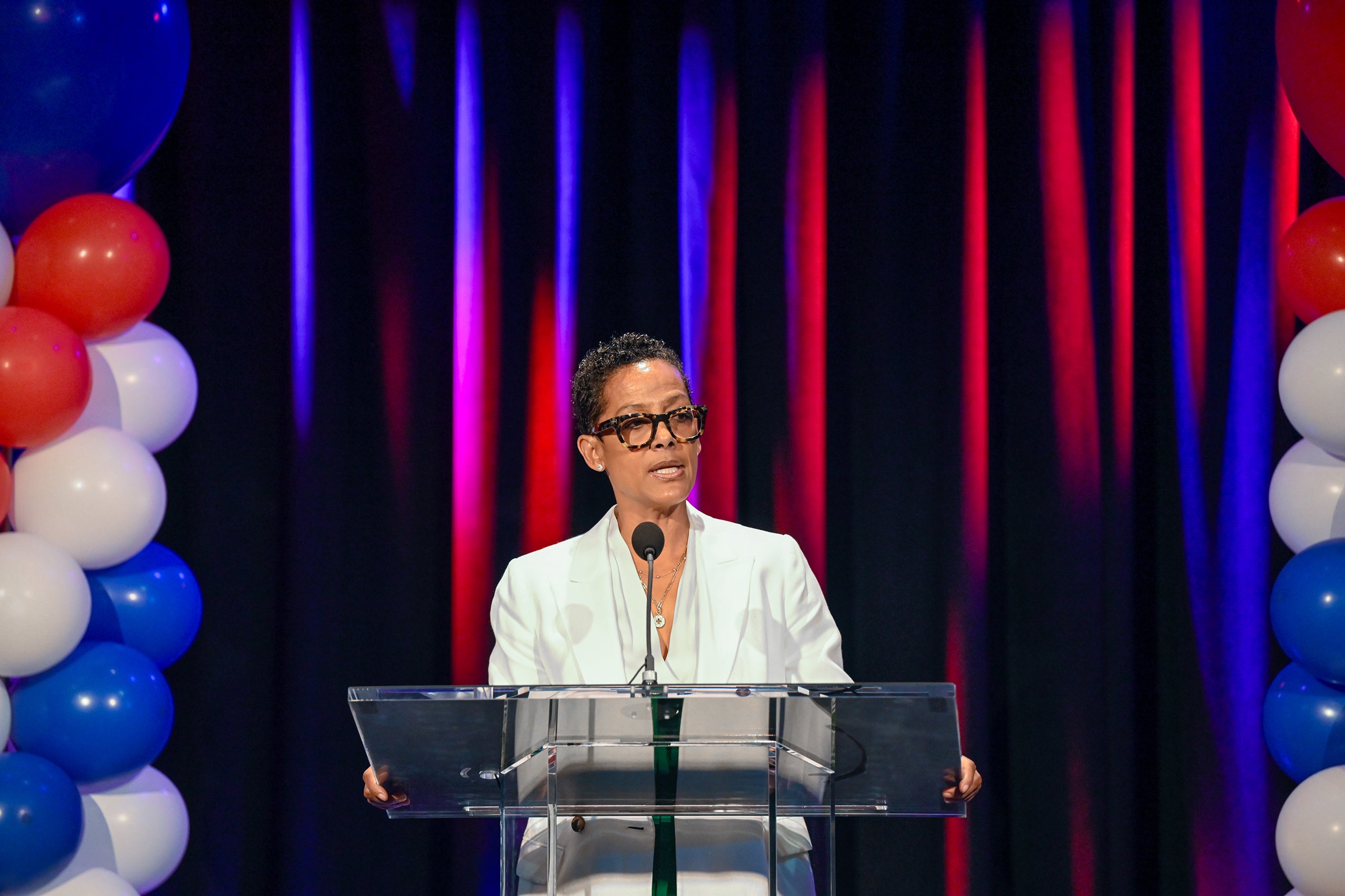 Renata Simril in a white suit stands behind a podium, speaking into a microphone, with a backdrop of black curtains and colorful lighting, flanked by red, white, and blue balloons on either side.