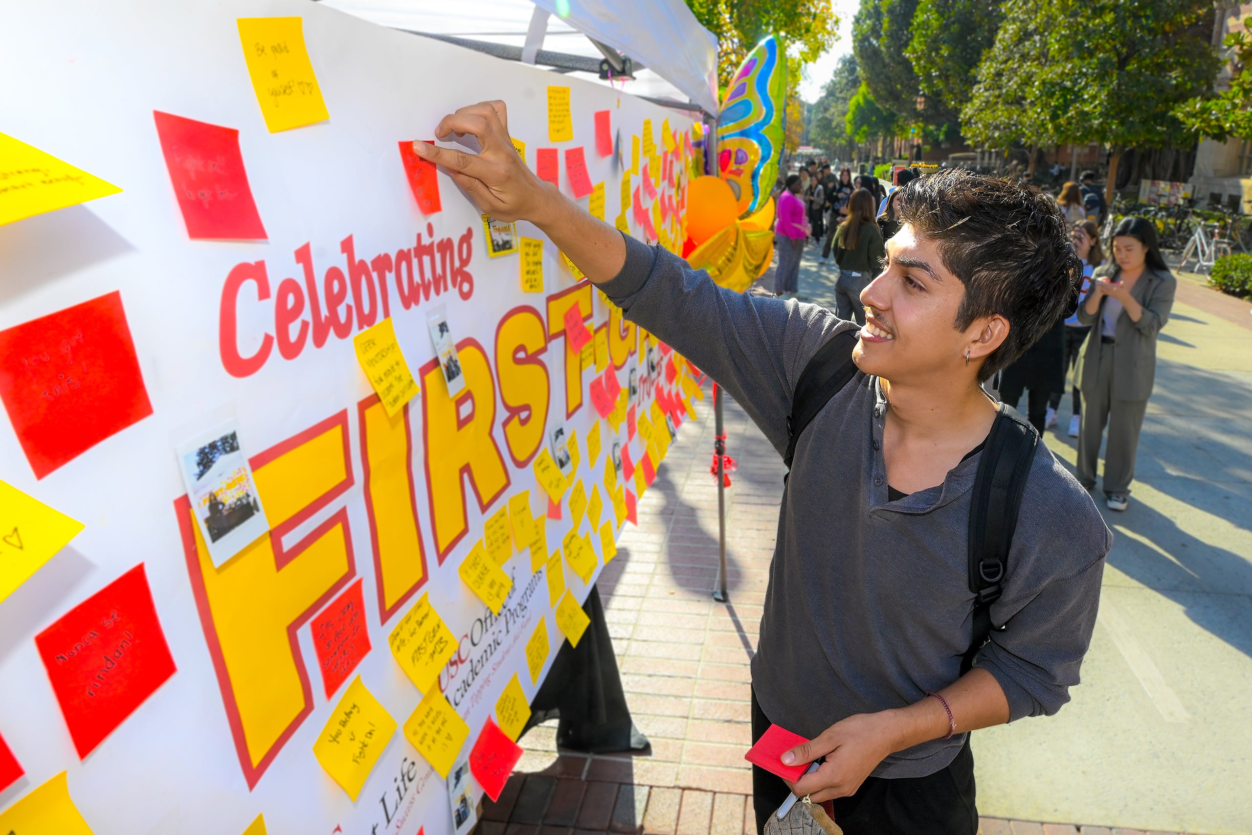 Person attaching a sticky note to a 'Celebrating FIRST' board with many notes, in an outdoor event setting.