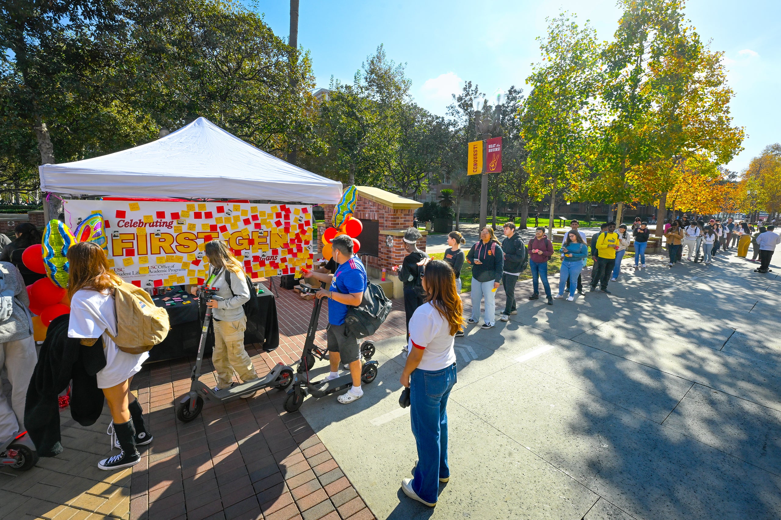 Outdoor event with 'Celebrating FIRST' booth, people, scooters, and a line forming in a sunny, tree-shaded area.