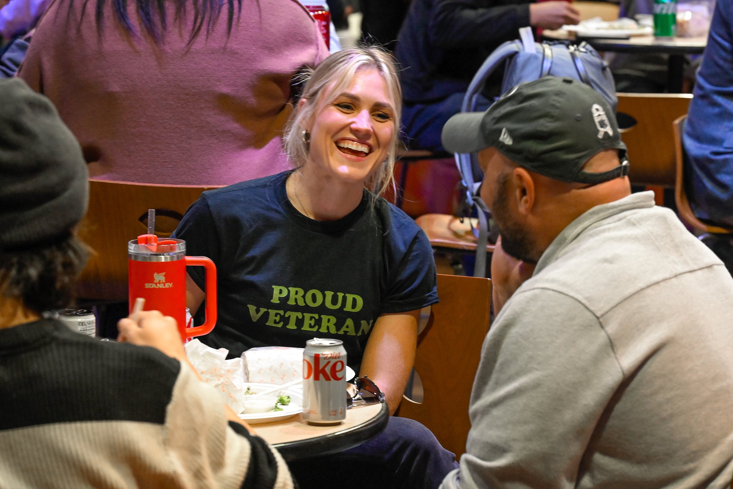 A person wearing a 'PROUD VETERAN' t-shirt sits with others at a table with drinks, in a casual dining setting.