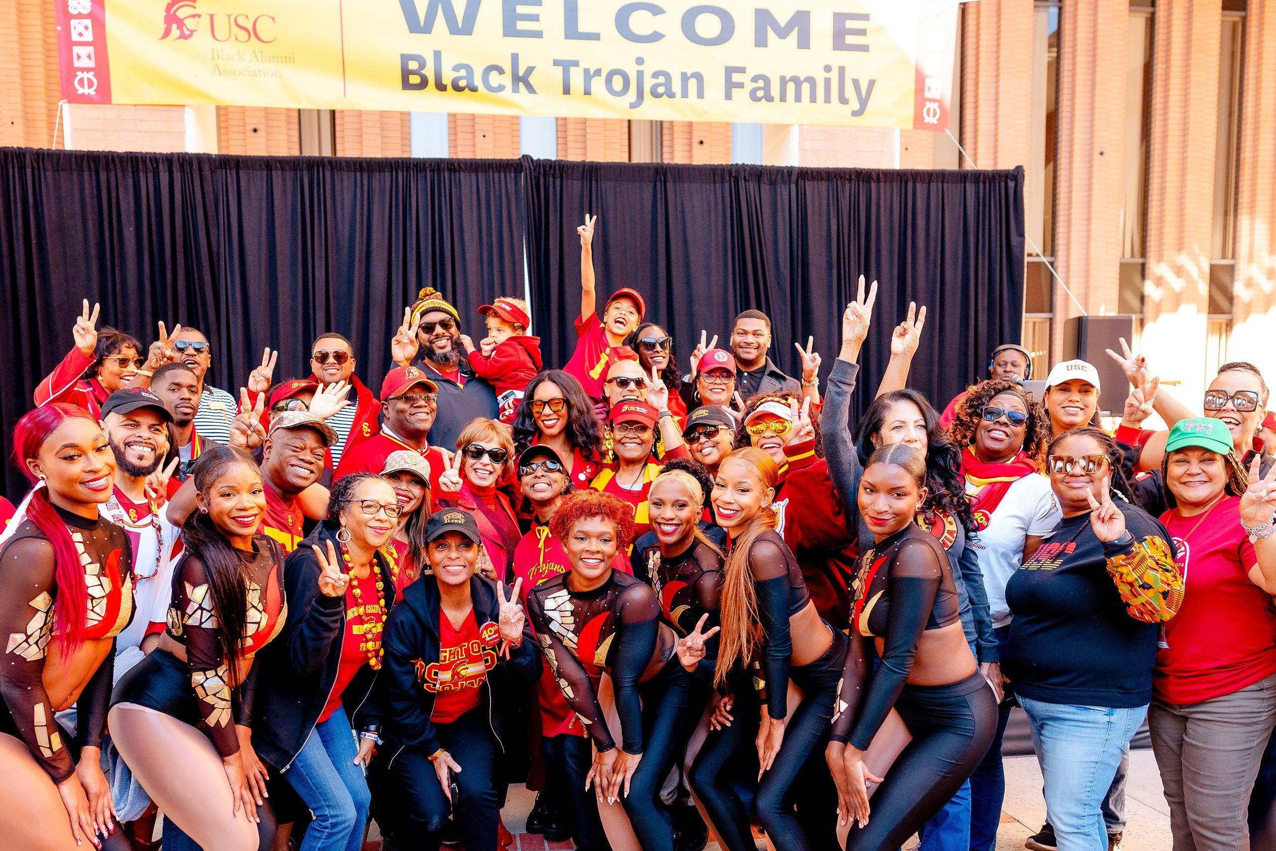  A large group of people, many wearing red and gold USC apparel, pose for a photo in front of a banner that reads 'Welcome Black Trojan Family,' with some individuals making Fight On signs.