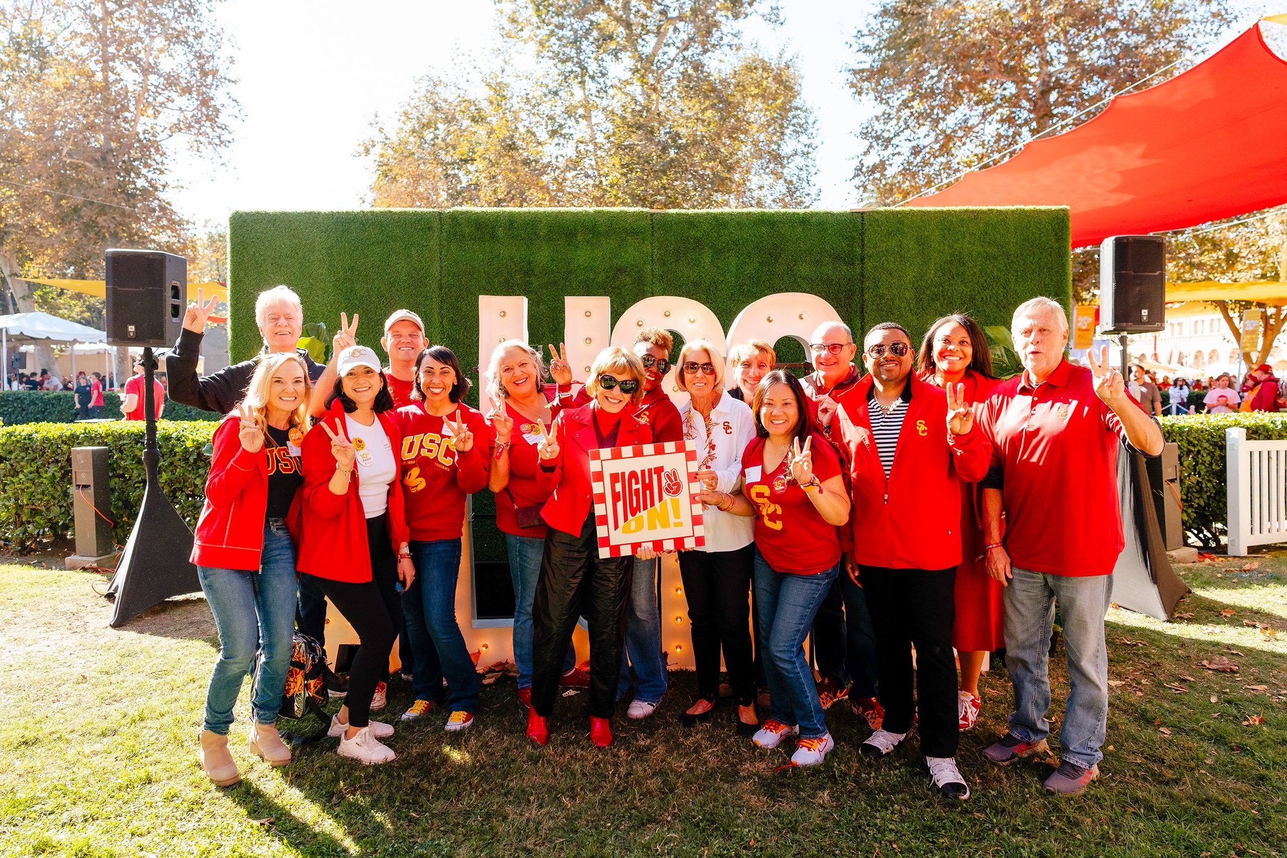 A group of people wearing red and gold, showing school spirit, pose for a photo in front of a large 'USC' sign, with President Carol Folt and others holding a 'Fight On!' sign.