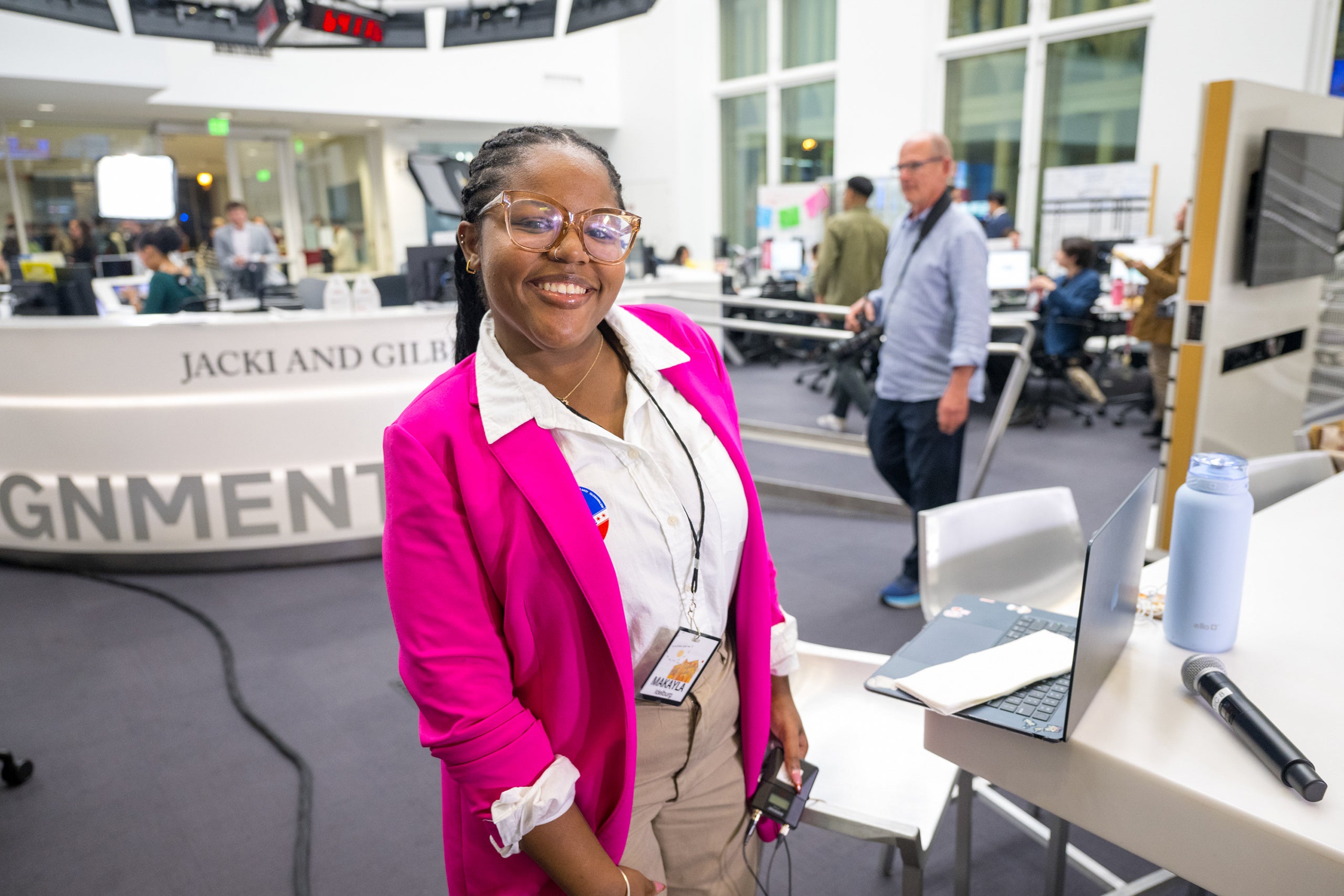 A person in a bright pink blazer and white shirt stands in a bustling newsroom environment with various people working in the background.