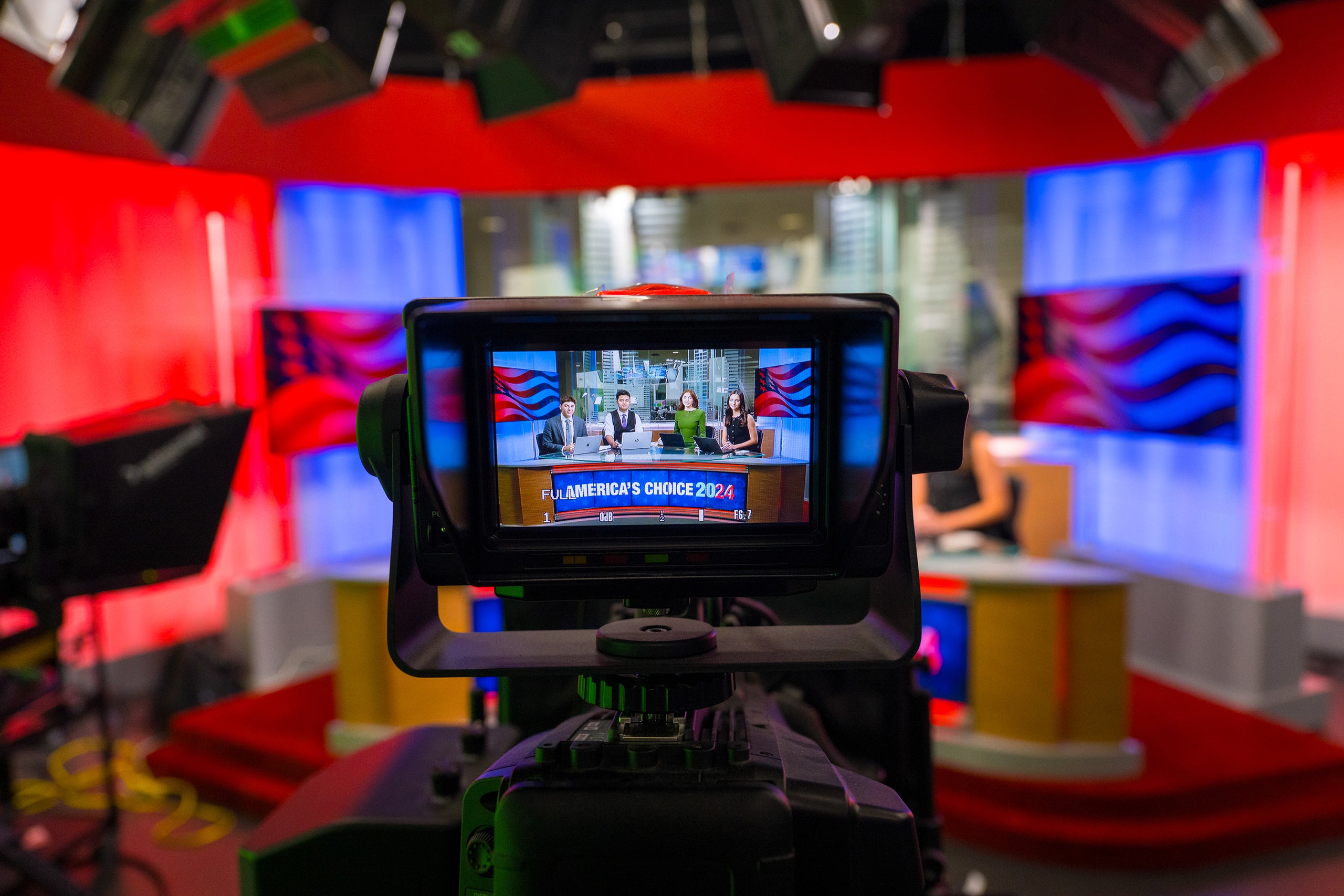 A camera view showing a news studio set with a large screen displaying a debate panel for 'America's Choice 2024.' The set is brightly lit with red and blue colors, and there are various pieces of studio equipment visible around.