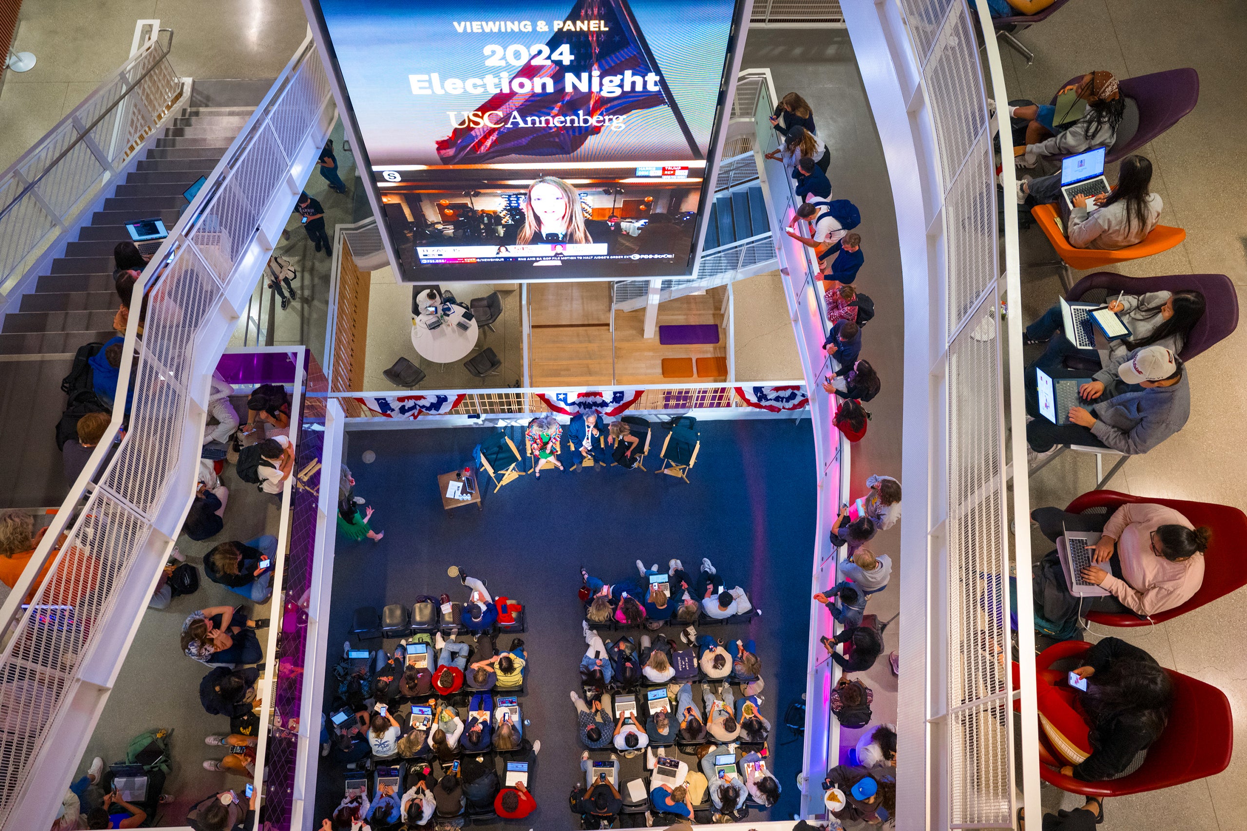 Overhead view of a crowded event with a screen showing '2024 Election Night,' people seated watching a panel, and others on laptops.