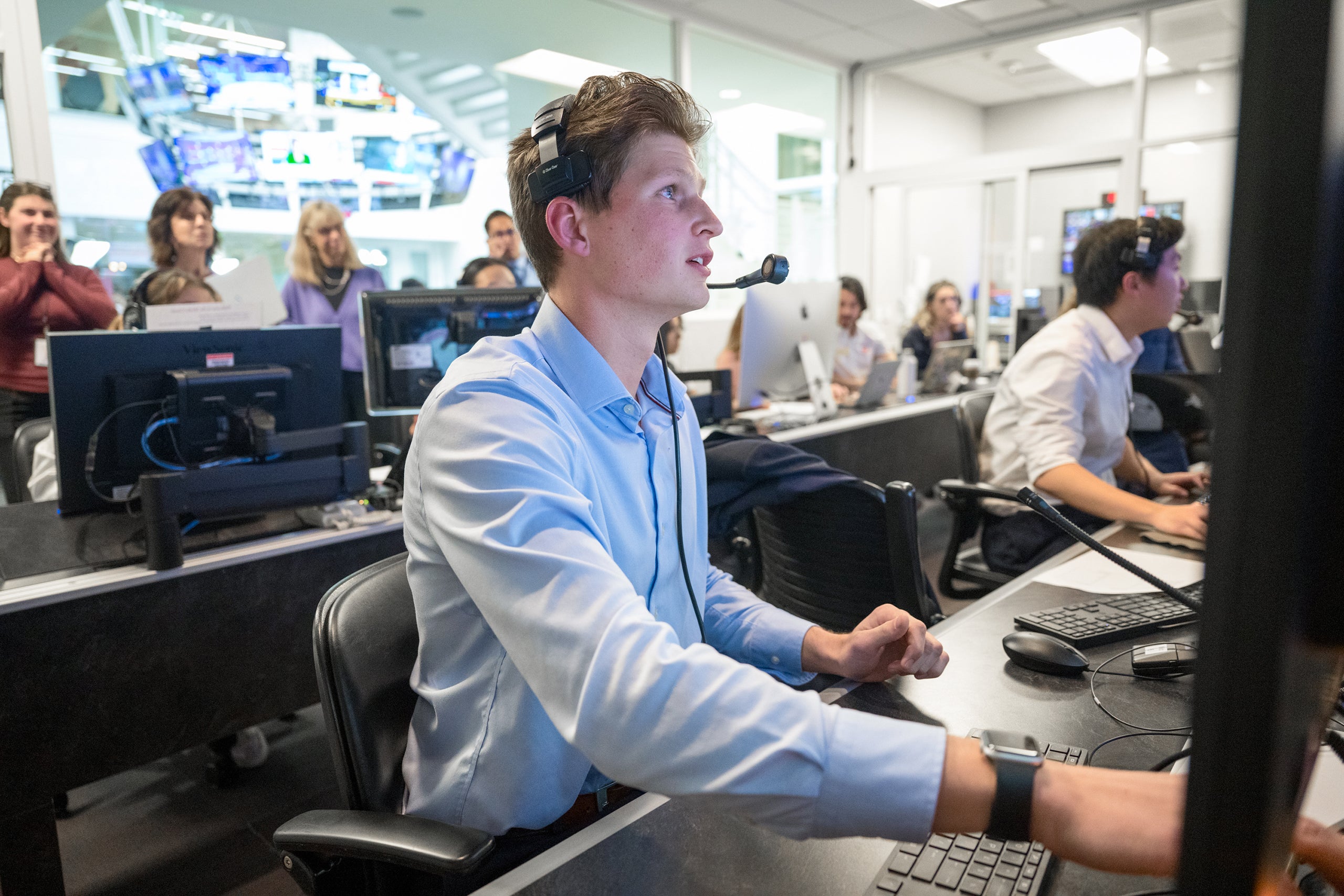 A man in a light blue shirt with a headset works at a control desk amidst multiple monitors in a busy broadcast room.