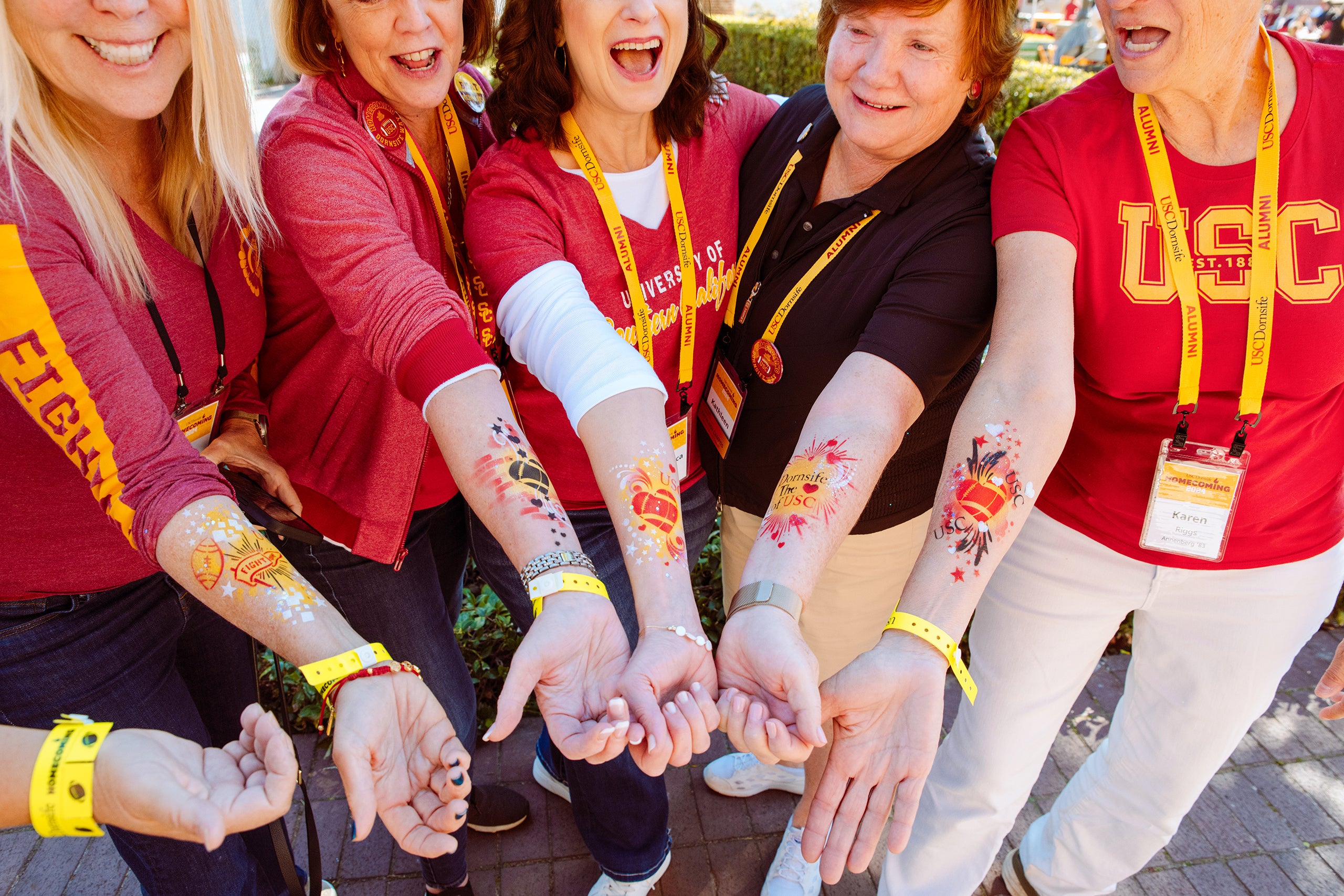 A group of people wearing red and gold USC apparel show their hands and forearms decorated with festive homecoming-themed paint, with yellow wristbands and lanyards indicating their participation in the event.